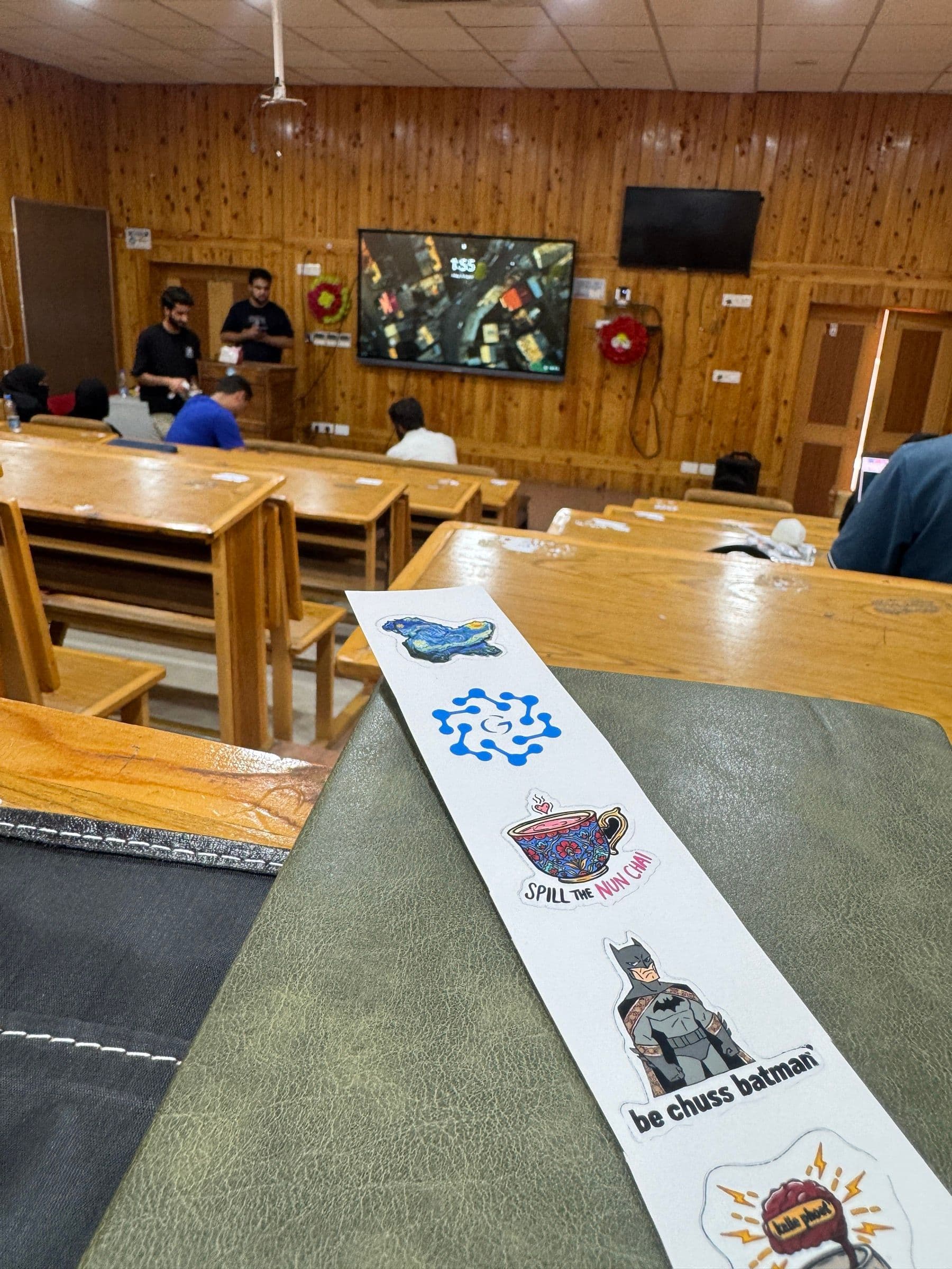 A strip of event stickers on a chair, including one reading 'be chuss batman' and a samovar captioned 'spill the nun chai'