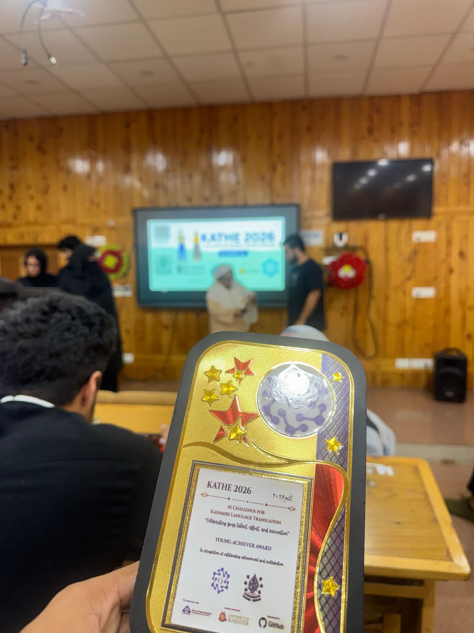 The KATHE 2026 Young Achiever Award plaque held up in the hall, the event slide on the screen behind it